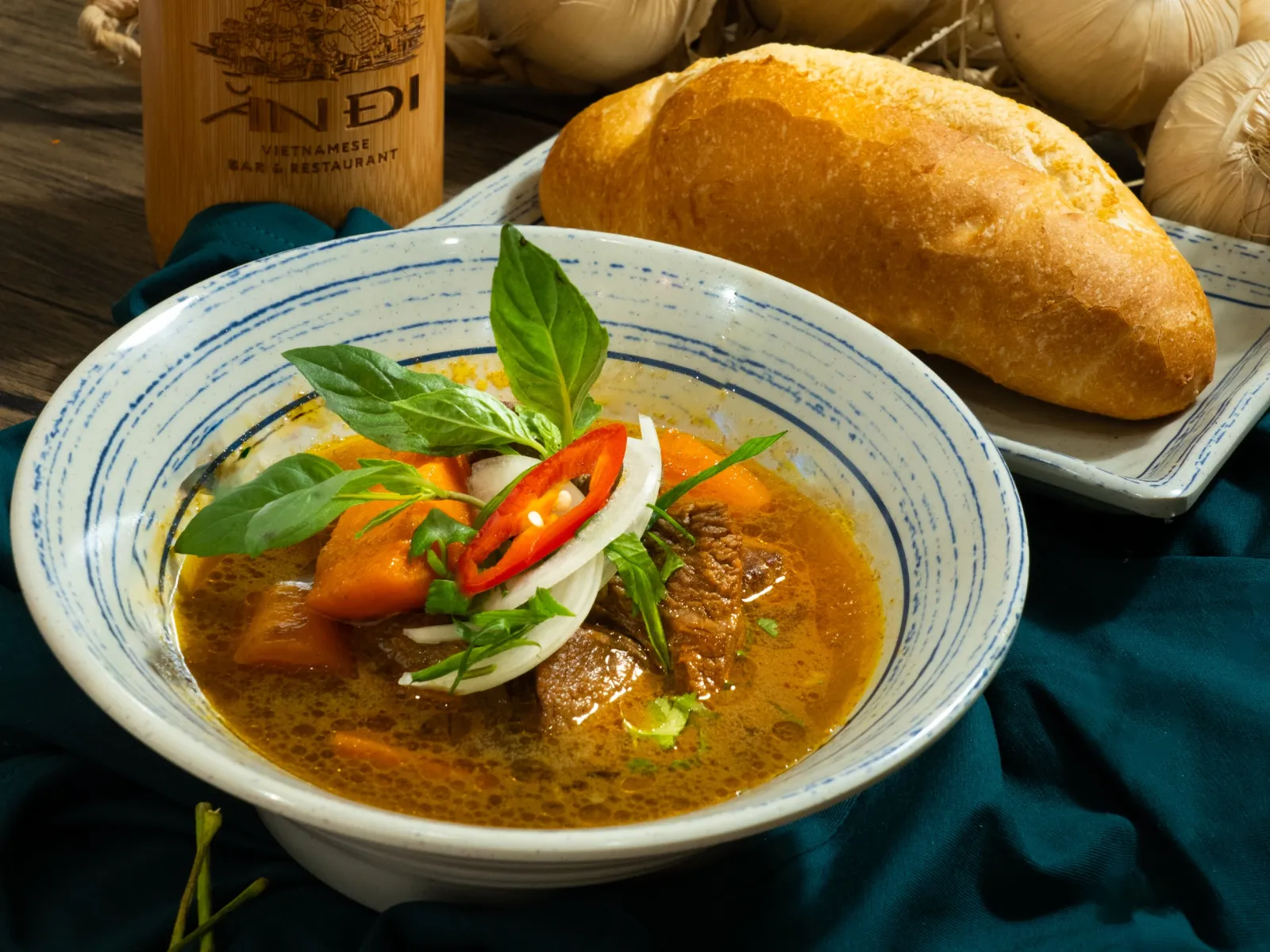 Bowl of bò kho beef stew with carrot, chilli and Thai basil, a bread roll behind
